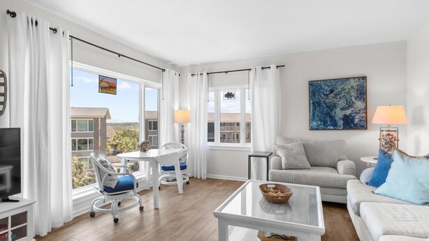 Un salon moderne avec un canapé blanc, une table basse en verre et un meuble pour la télévision. On y trouve deux fauteuils avec des coussins bleus, une petite table avec une lampe et un tableau accroché au mur.