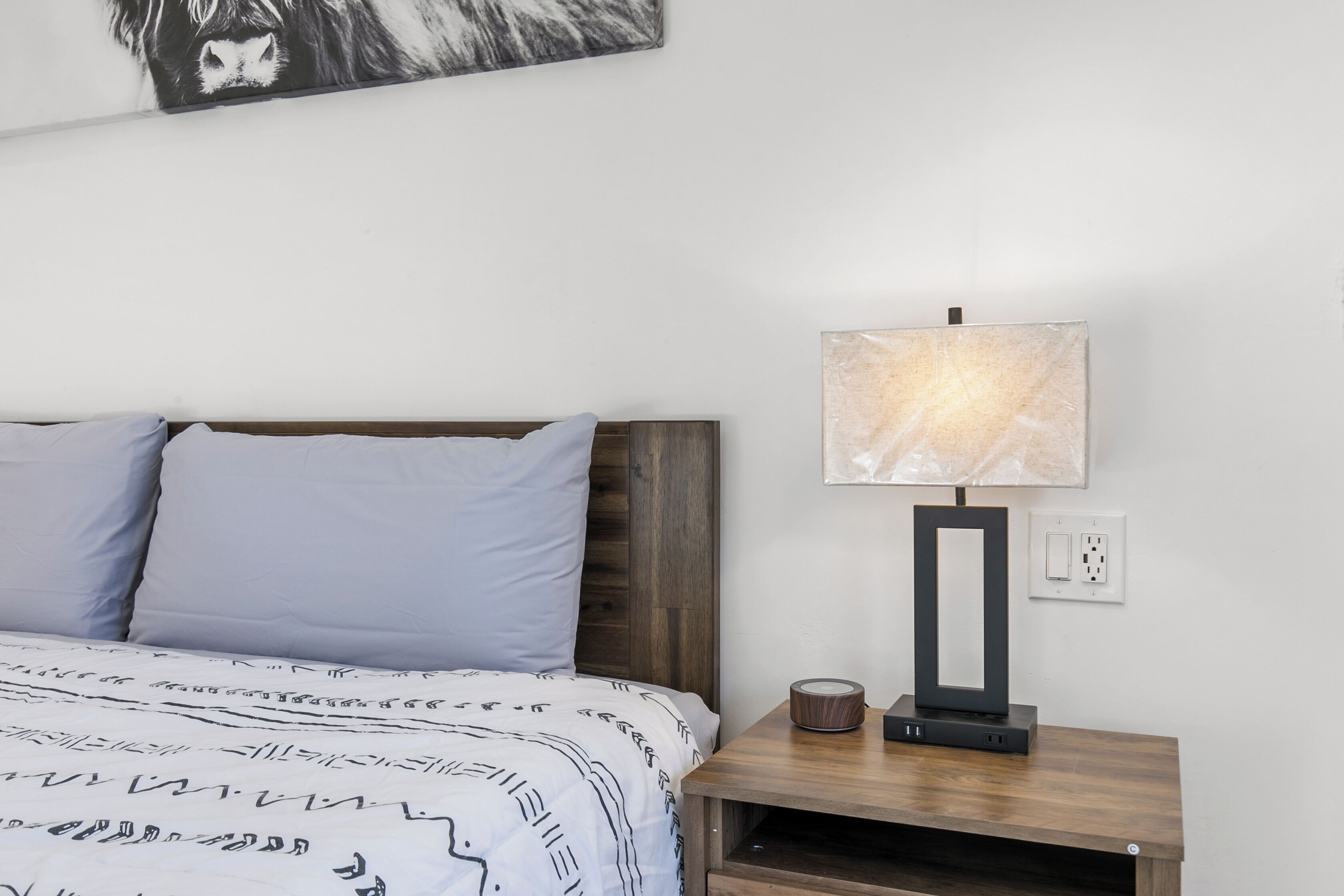 A bedroom with a bed, a nightstand with a lamp, and a wall-mounted power outlet.