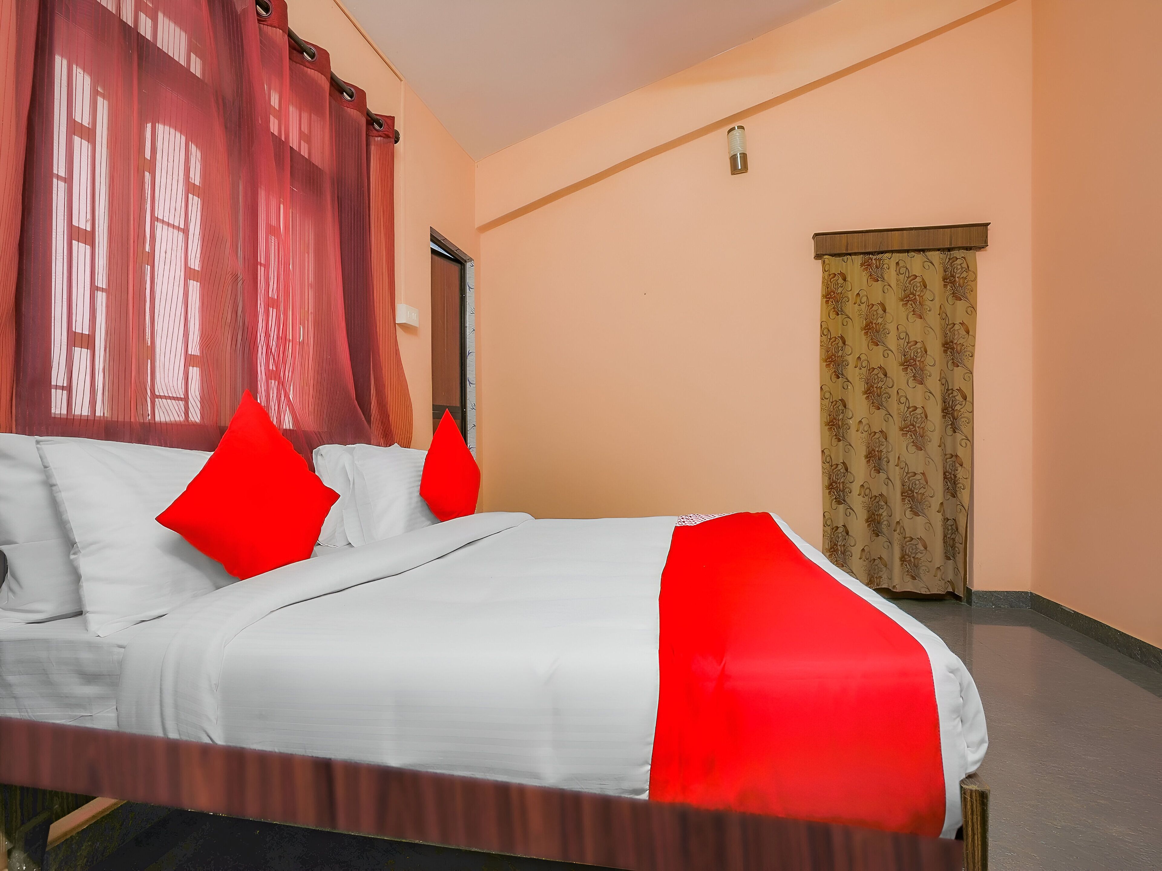 A hotel room with a bed, red and white bedding, a wooden headboard, a door, and a patterned curtain.