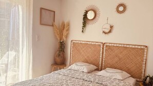 A bedroom with a large bed, a window with sheer curtains, a bedside table with a vase of pampas grass, and two wall-mounted decorative mirrors.