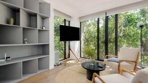 A modern living room with a large TV, a round coffee table, and a comfortable chair. There are shelves with decorative items and a view of trees through large windows. - Breathtaking loft in Roma Norte (Ciudad de México)