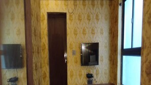 Ein Zimmer mit Holztür, einem an der Wand montierten kleinen Fernseher, einem Fenster und einer hölzernen Bank.