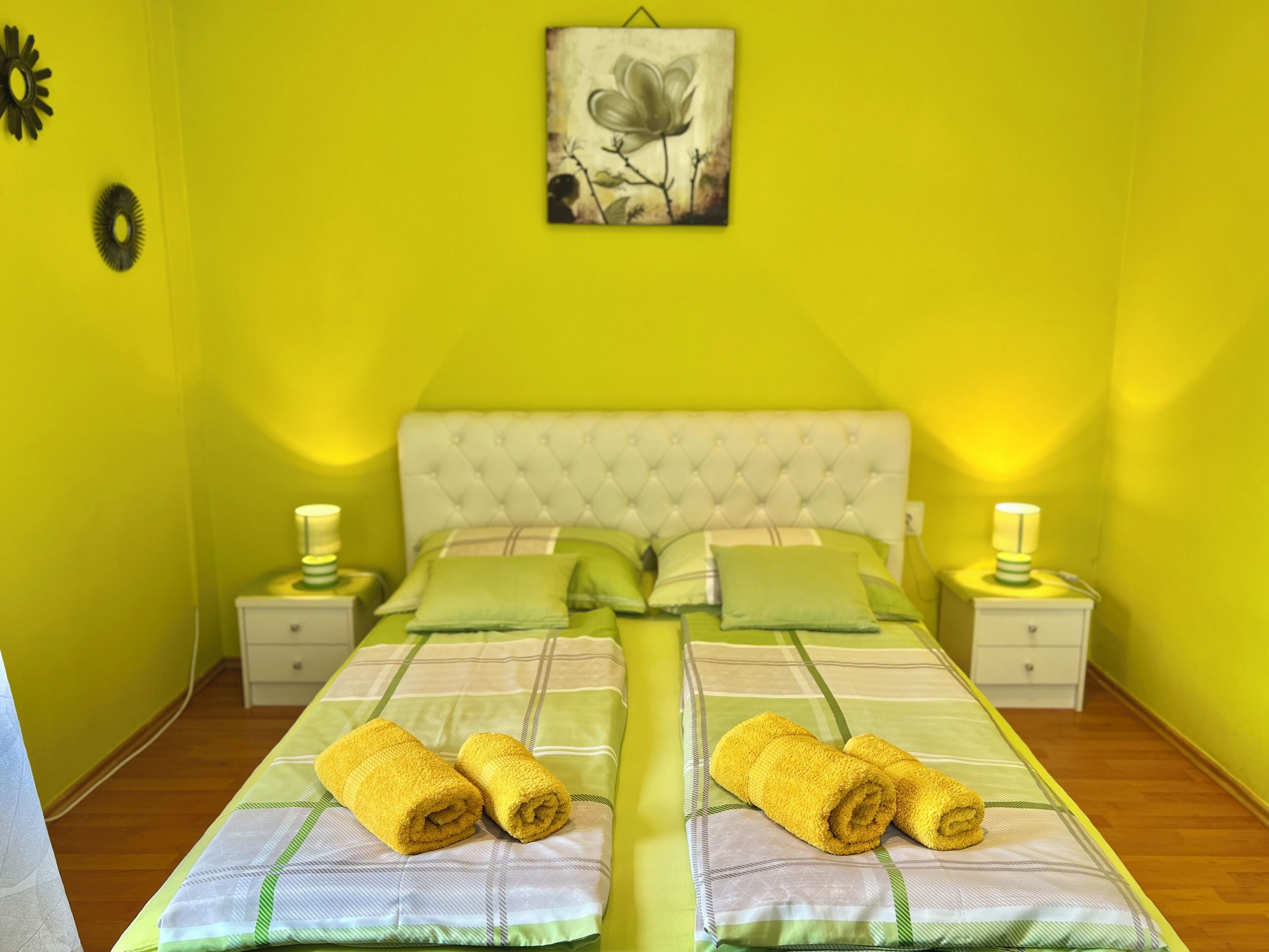 A double bed with a tufted headboard, two nightstands with lamps, green and white bedding, and a wall art piece above the bed.