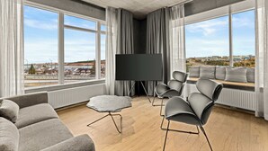 Una sala de estar moderna con un televisor grande, sofás grises, una mesa de centro redonda y vistas de la ciudad a través de amplias ventanas.