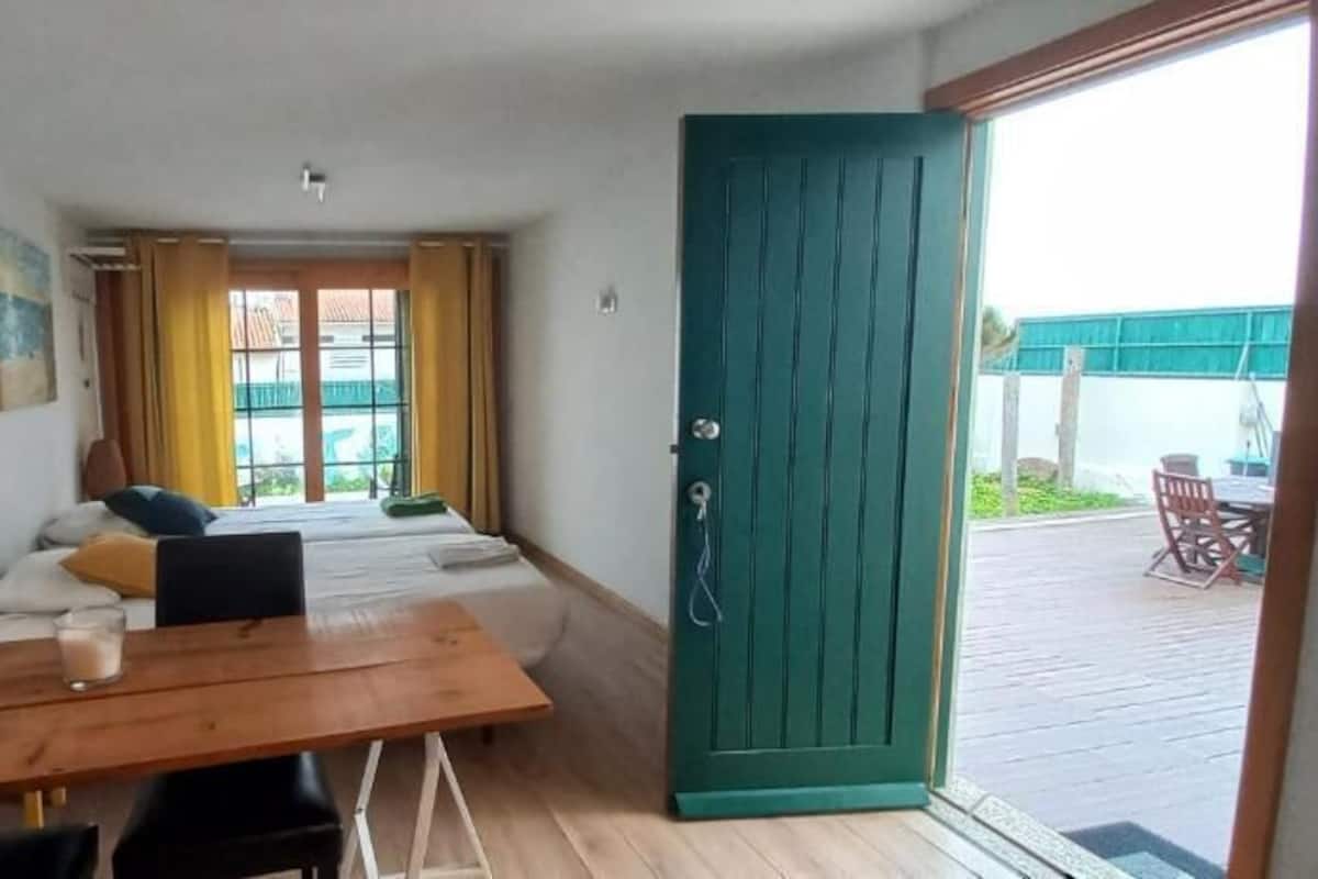 Une pièce comprenant un lit, une table avec des chaises, une porte verte et donnant sur un espace extérieur aménagé avec une table et des chaises.