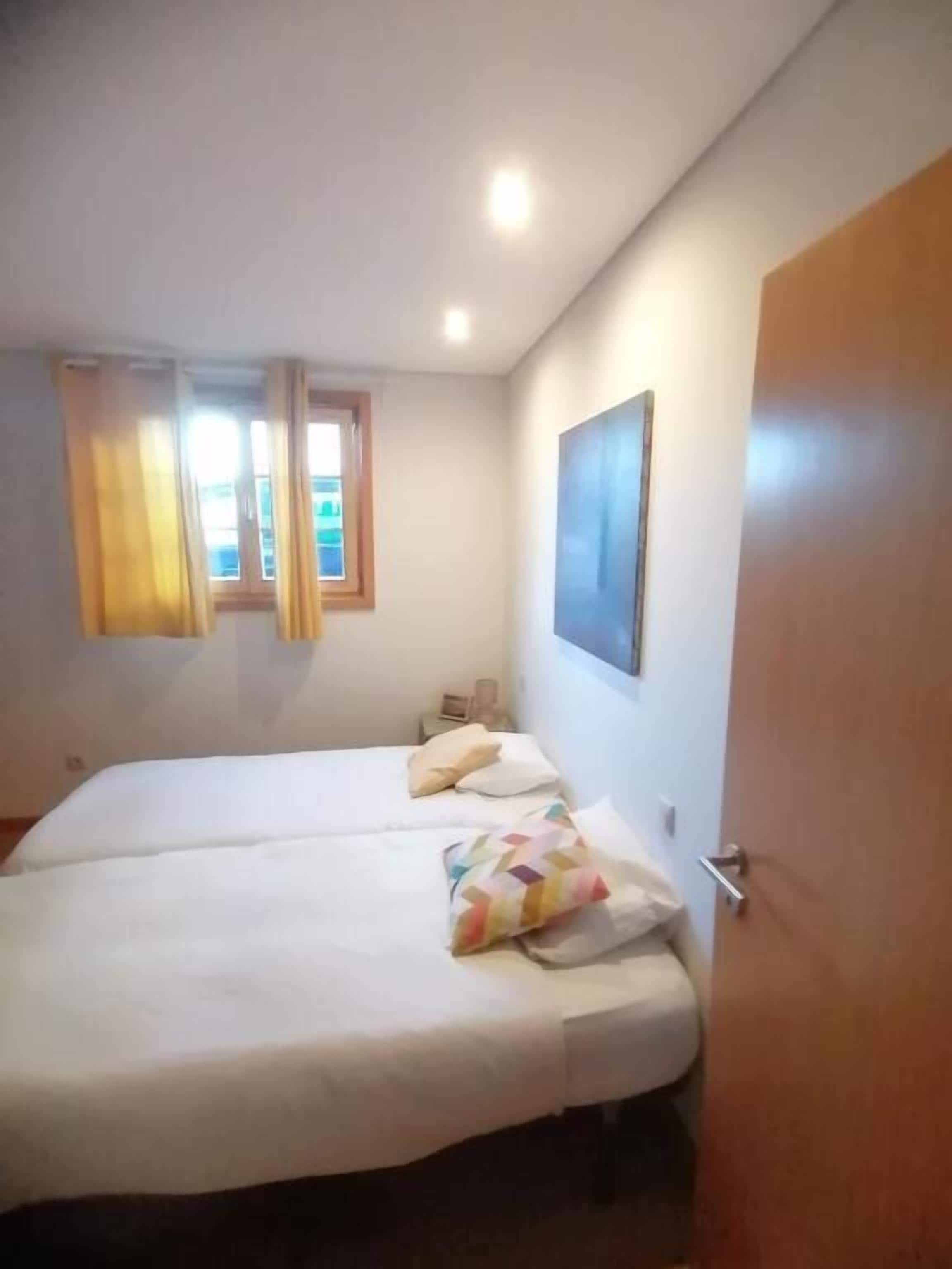 Une chambre à coucher avec un lit double, une fenêtre avec des rideaux, une table de chevet et une porte.