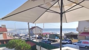 Un balcón con una mesa y sillas de madera, una sombrilla y vistas al océano y coches estacionados.