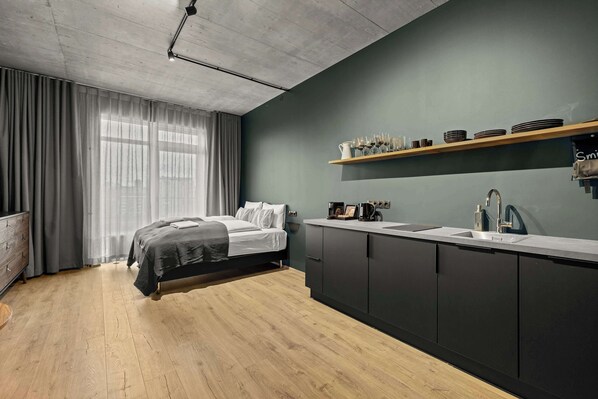 A modern bedroom with a bed, a large window with curtains, a wooden floor, a kitchen area with a sink, and a shelf with glasses and plates.
