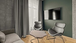 Ein modernes Wohnzimmer mit grauem Sofa, zwei grauen Sesseln, einem kleinen runden Tisch, einem Flachbildfernseher, der an einer grünen Wand befestigt ist, und einem großen Fenster mit grauen Vorhängen.