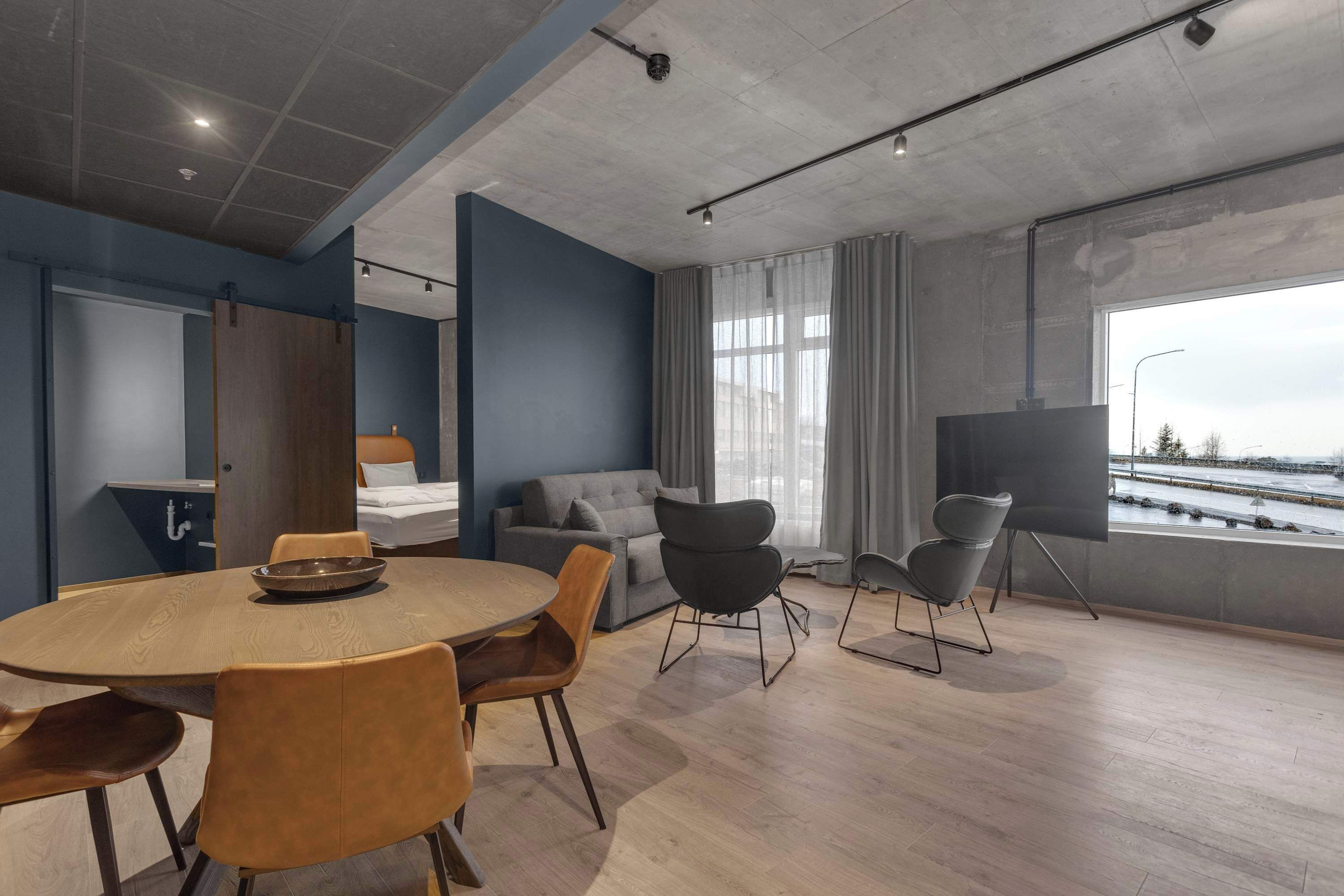 A modern hotel room with a round wooden dining table, comfortable seating, a grey sofa, a large window with a view, and a flat-screen TV.
