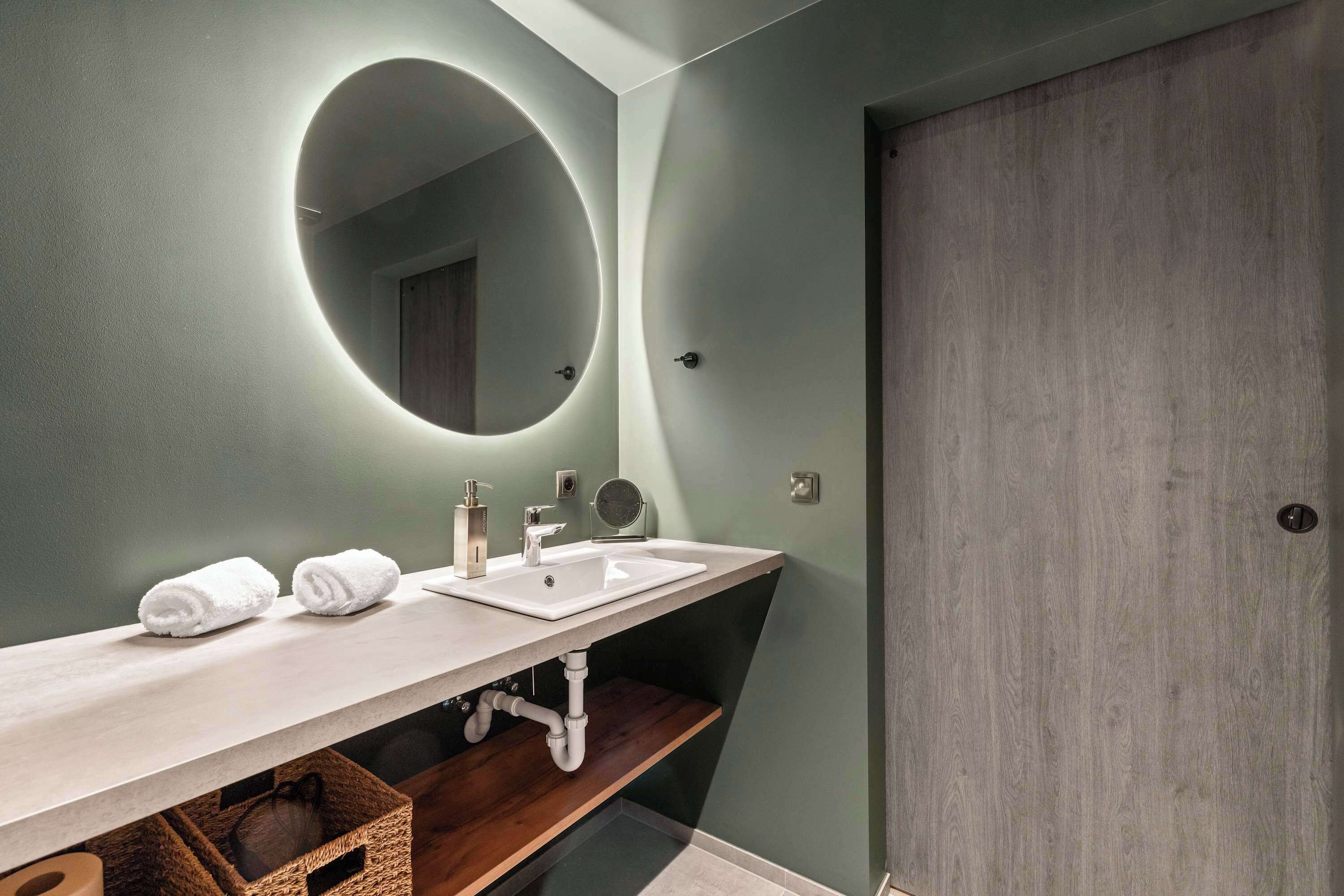A modern bathroom with a double sink, a round mirror, and a wooden door.