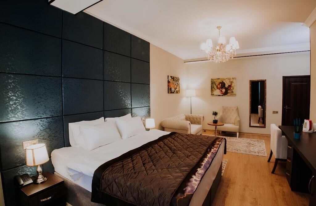 A bedroom with a large bed, a nightstand with a lamp, a chair, a desk, and a chandelier.