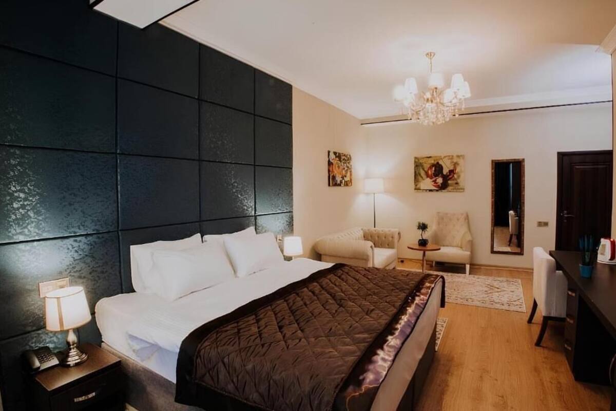 A bedroom with a large bed, a nightstand with a lamp, a chair, a desk, and a chandelier.