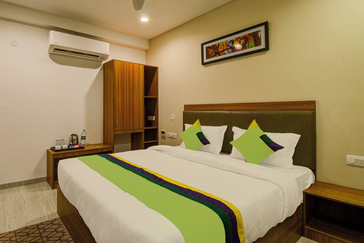 A hotel room with a large bed, a wooden wardrobe, a bedside table with a lamp, and an air conditioning unit.