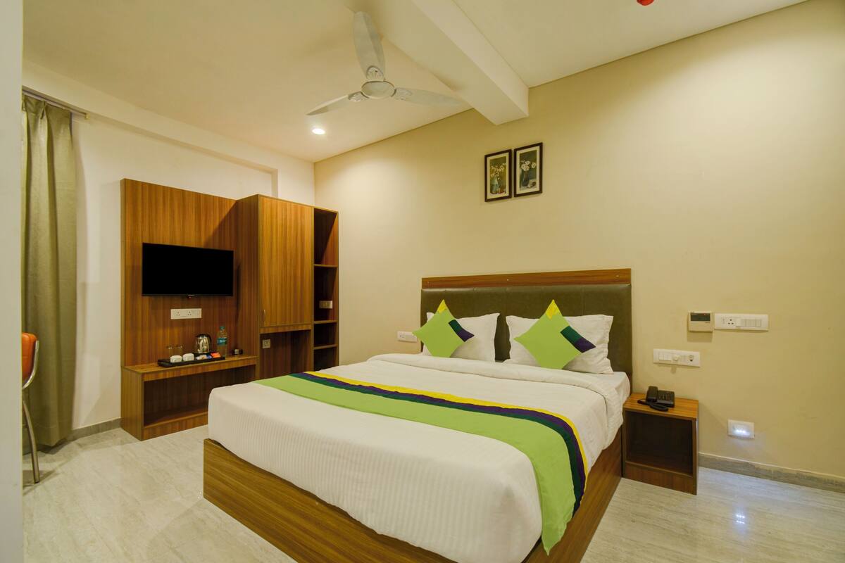 A hotel room with a bed, a TV mounted on the wall, a wooden wardrobe, a bedside table, and a ceiling fan.