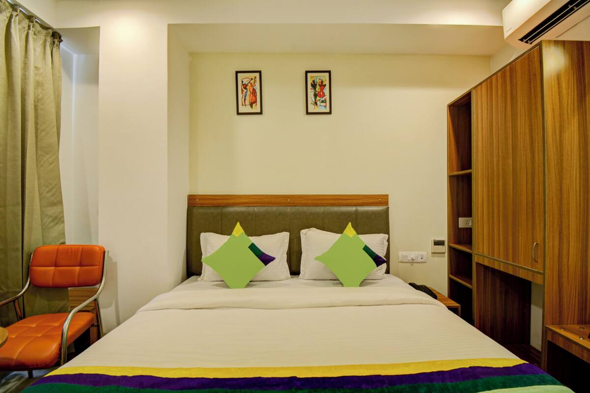 A hotel room with a bed, two pillows, a chair, a wardrobe, and two framed pictures on the wall.