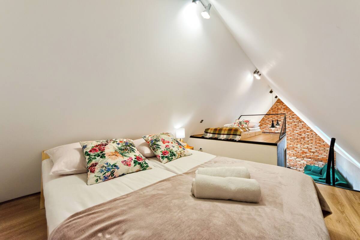 Un dormitorio de estilo loft, con una cama con almohadas florales, cabecera de madera y vista a una pared de ladrillo a través de una ventana abierta.