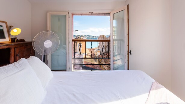 Ein Hotelzimmer mit Bett, Standventilator, Nachttisch mit Lampe und Blick auf eine Stadt durch eine offene Tür.