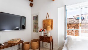 Ein gemütliches Zimmer mit Fernseher, einem Holztischchen, einem Korbwäscheschrank, einer Lampe und Blick auf ein Backsteingebäude durch ein Fenster.