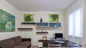 A dining area with a brown table, four chairs, and a decorative plate. There are two shelves above the table, one with a vase and the other with a small plant. A brown sofa is against the wall. - FIORDALISi Apartment (Desenzano del Garda)