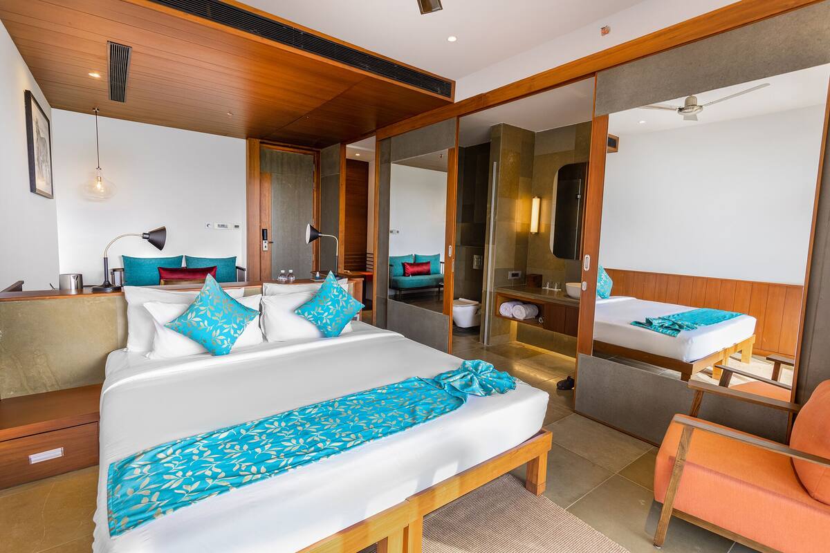 A modern hotel room with a large bed, a wooden bench, a bathroom with a glass shower, and a seating area with a chair.