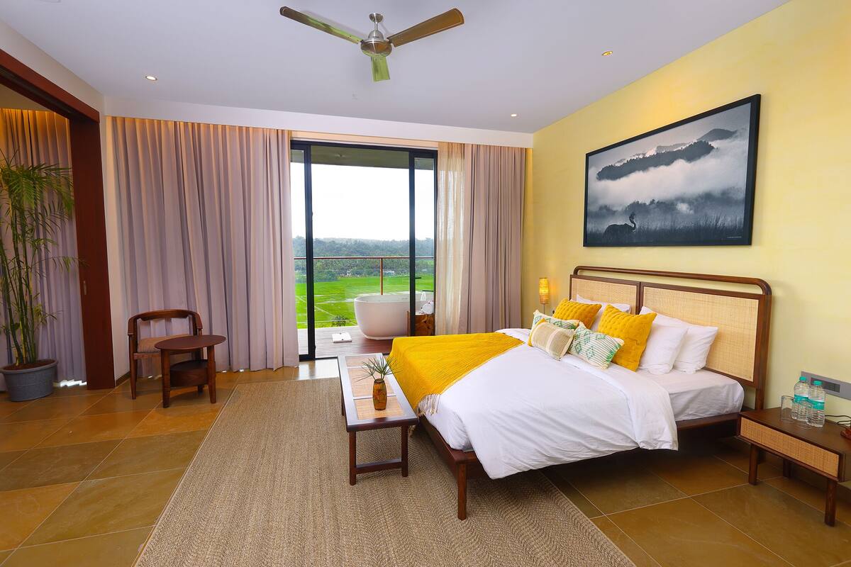 A spacious bedroom with a large bed, a view of the outdoors, a ceiling fan, and a painting on the wall.