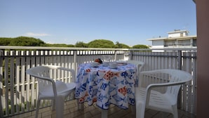 A balcony with a table set for two, white plastic chairs, and a view of the sea and buildings in the background. - Contemporary apartment in the heart of Lido del Sole (San Michele Al Tagliamento)
