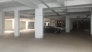 An underground parking garage with concrete pillars, a few parked cars, and a visible exit door. - Hotel White House (Ahmedabad)