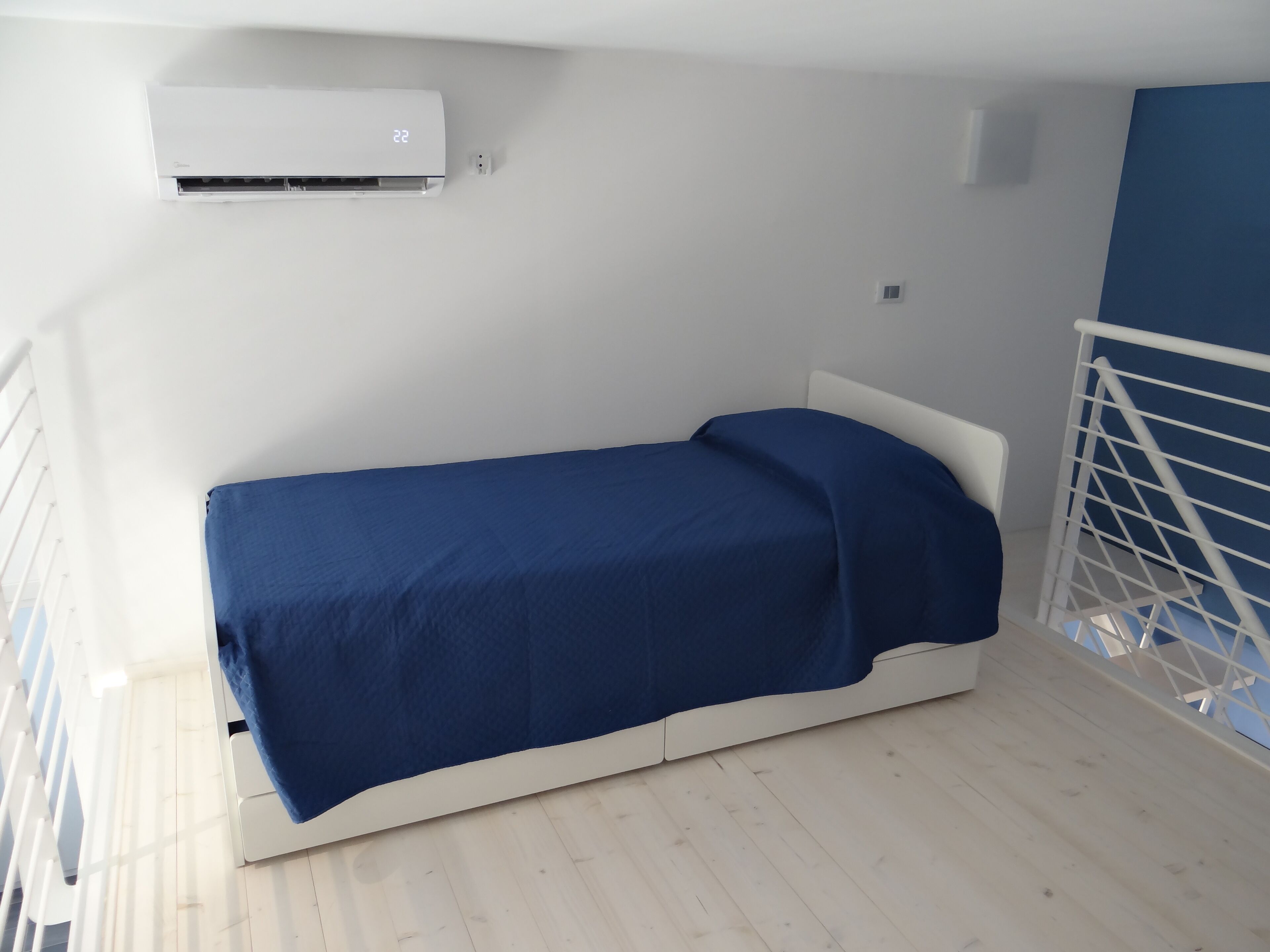 A small, single-bed room with a white headboard, a blue bedspread, and a white air conditioning unit mounted on the wall.