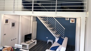 A modern living space with a white sofa, a TV on a white stand, a small table with snacks, and a staircase leading to an upper level.