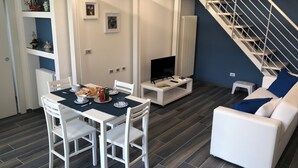 A modern living room with a dining area, white furniture, a TV, and a staircase.