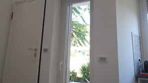 A room with a door, a window with blinds, and a view of greenery outside.