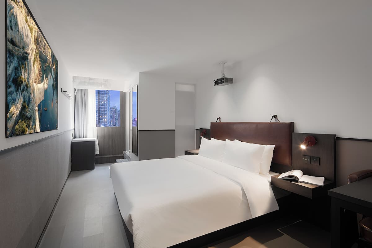 A modern hotel room with a large bed, a nightstand, a desk, and a large window offering a city view.