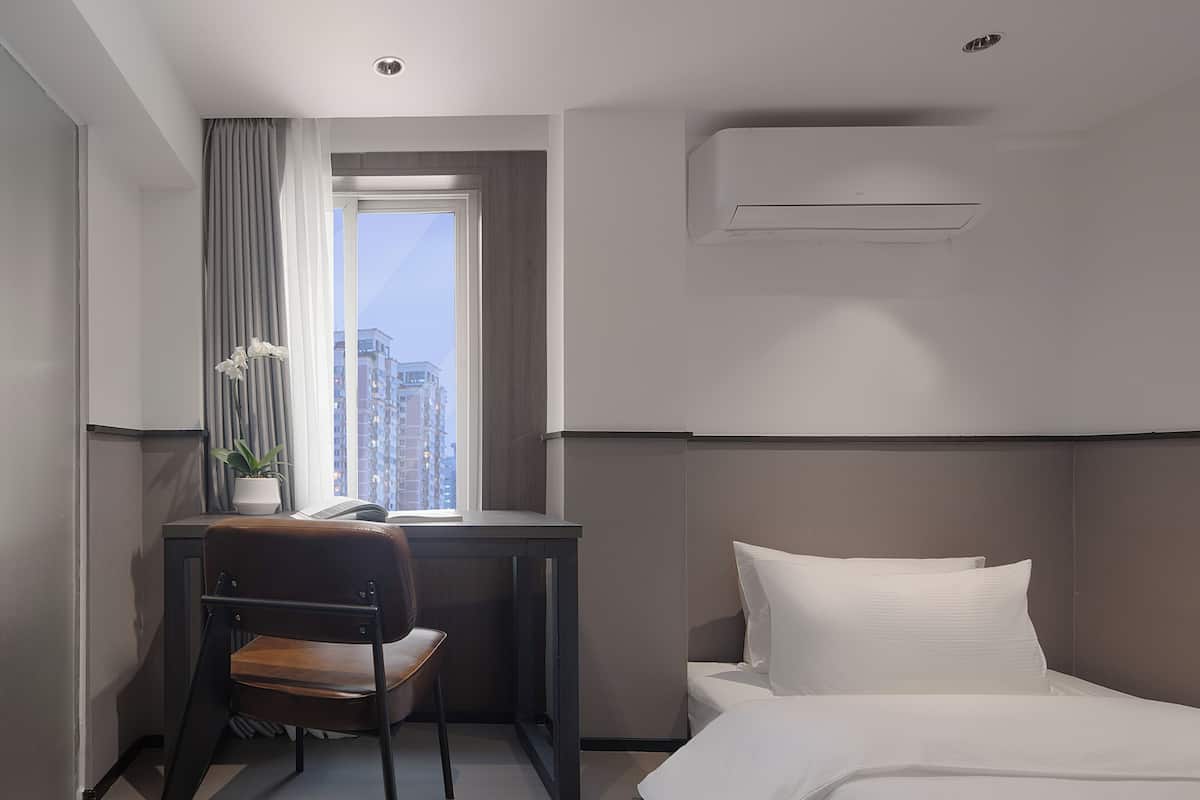 A hotel room with a bed, a desk, a chair, a potted plant, and a window with curtains.