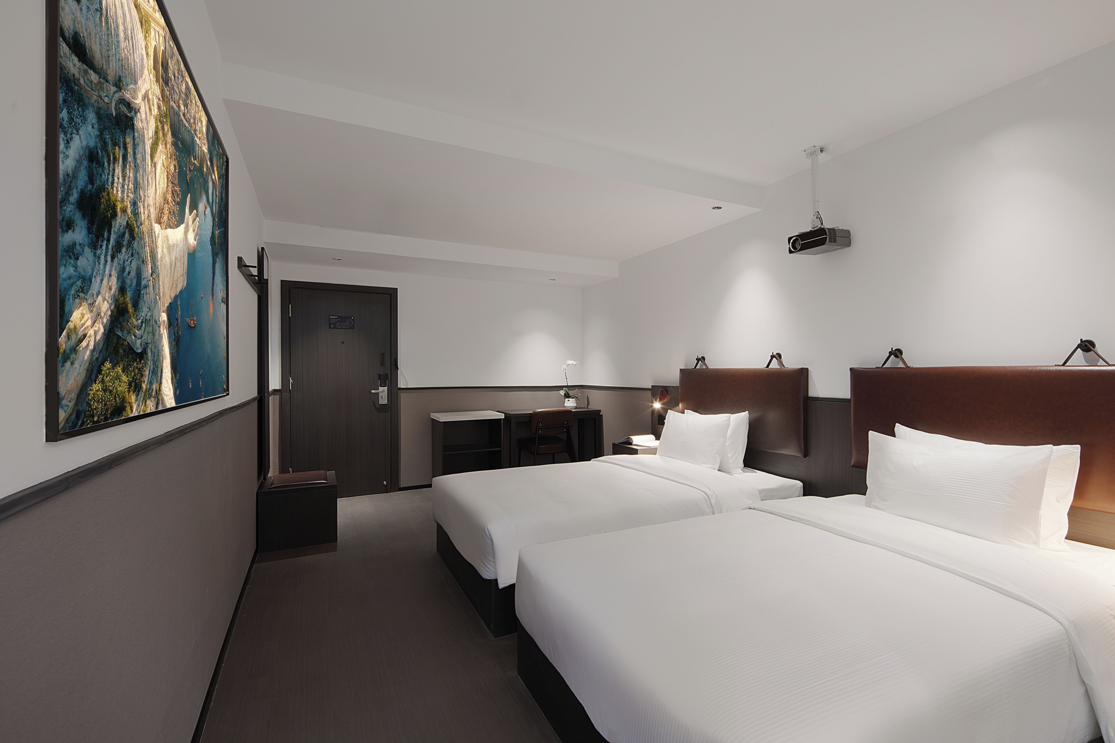 Habitación de hotel con dos camas, un escritorio, una silla, una mesita de noche y un cuadro grande en la pared.