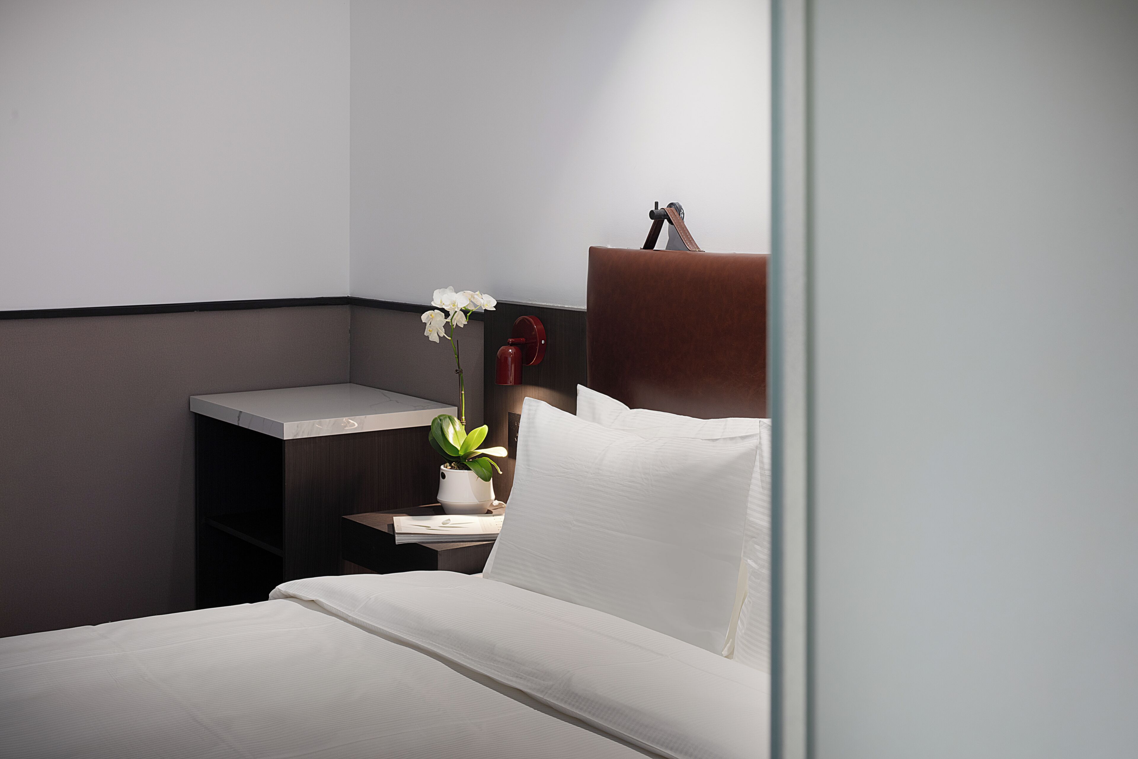 Una habitación de hotel con una cama, una mesita de noche, una planta en maceta y un objeto rojo sobre la mesita.