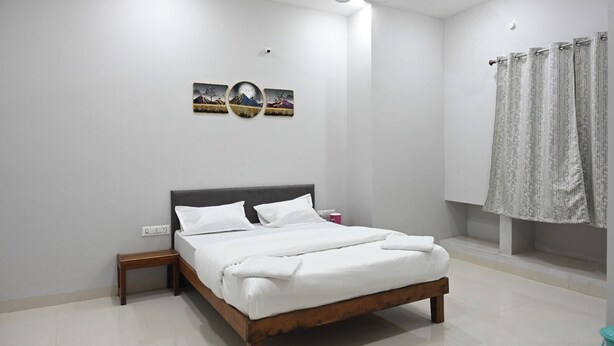 A bedroom with a wooden bed, white bedding, a nightstand, a wall-mounted light, and a window with curtains. - Hotel RRR Palace (Maheshwar)