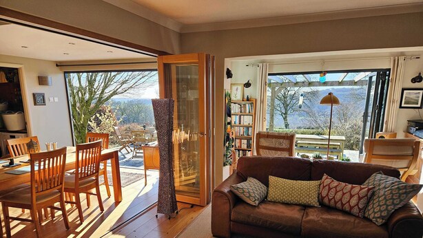 A cozy living room with a wooden dining table, chairs, a brown sofa with patterned cushions, a coffee table, and a bookshelf. - Couples retreat on Dartmoor with stunning views (Mary Tavy)