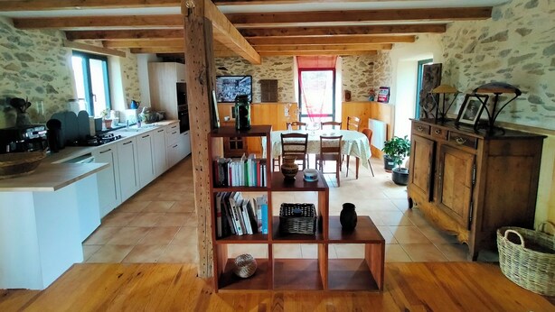 A rustic kitchen with wooden beams, a dining area with a table and chairs, and a bookshelf filled with books and decorative items. - A small house above the meadows. Light, space and silence. (Rieupeyroux)