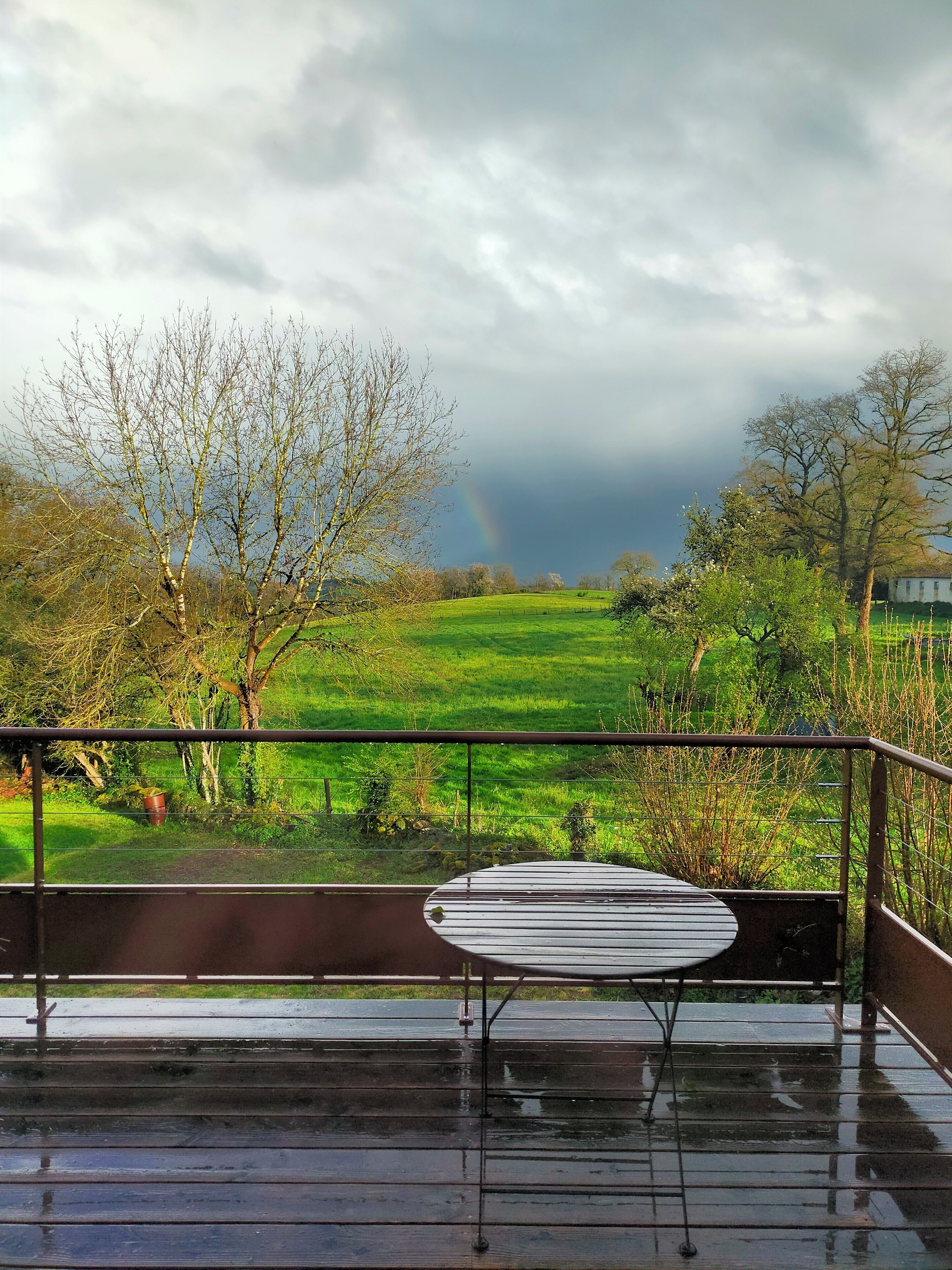 A balcony with a metal table and chairs, overlooking a green field with trees and a rainbow in the distance.
