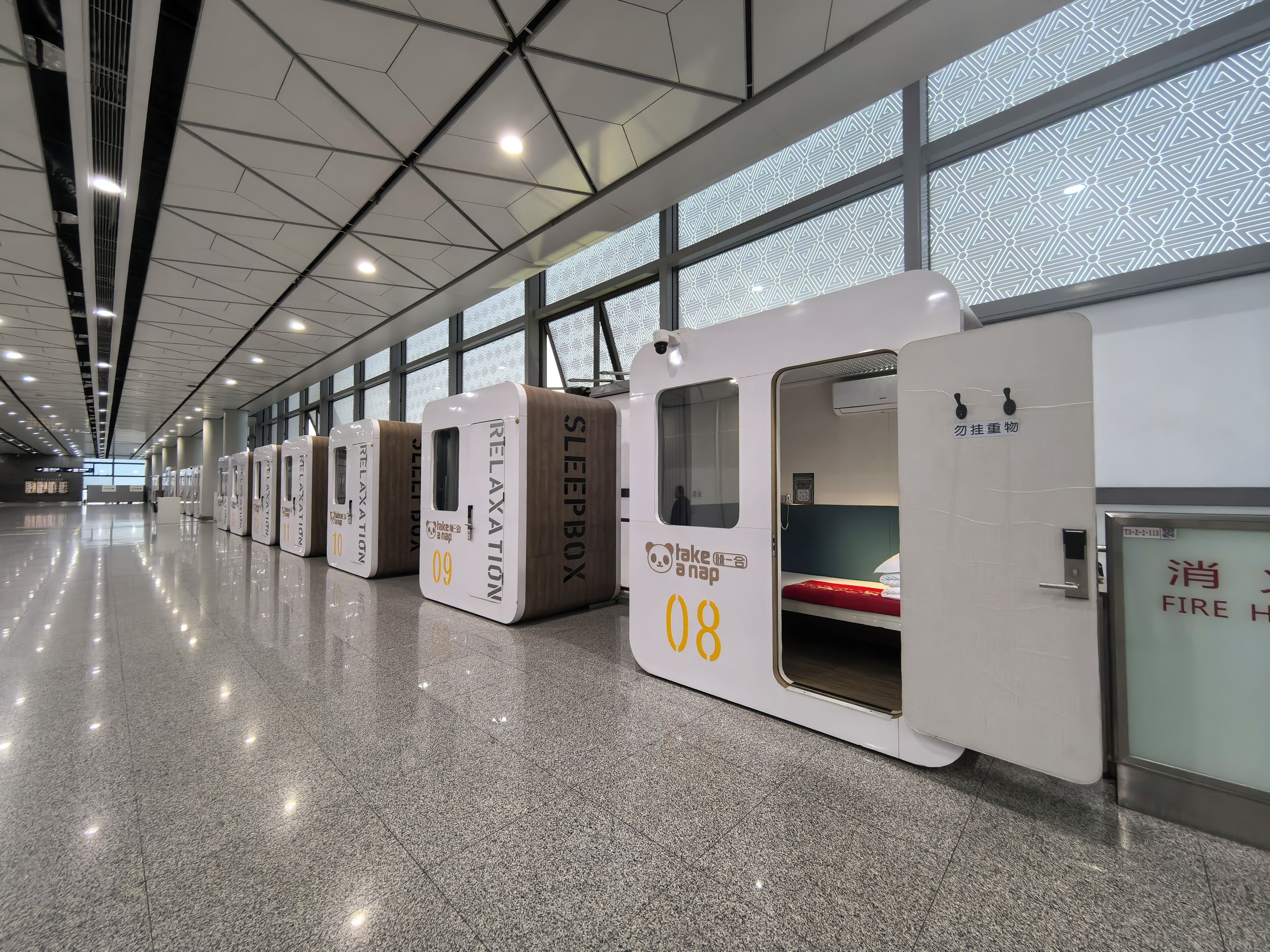 Una fila de cápsulas para dormir con los números 08, 09 y 10, en un entorno interior moderno con piso pulido y ventanas amplias.