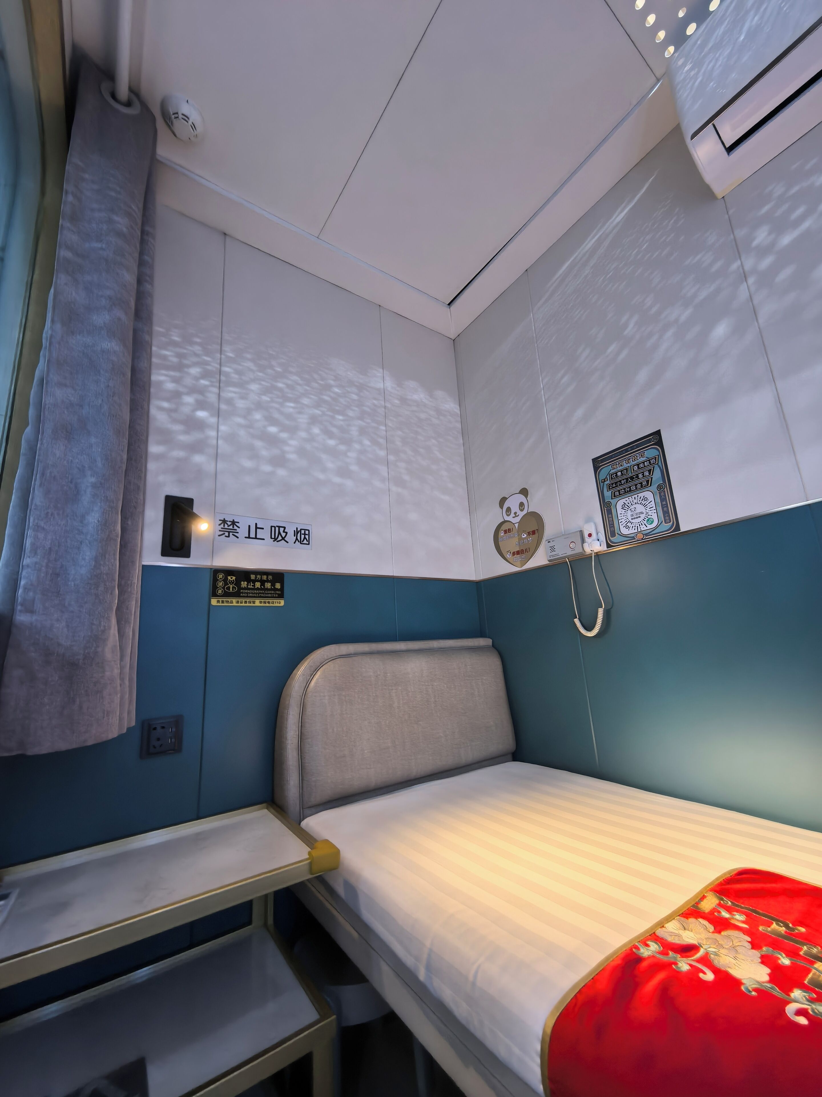 A compact train compartment with a single bed, a small table, and a wall-mounted air conditioning unit.