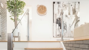 Un escalier avec des marches en bois et une rampe en verre, menant à un palier agrémenté d’une plante et d’une œuvre d’art abstraite au mur.