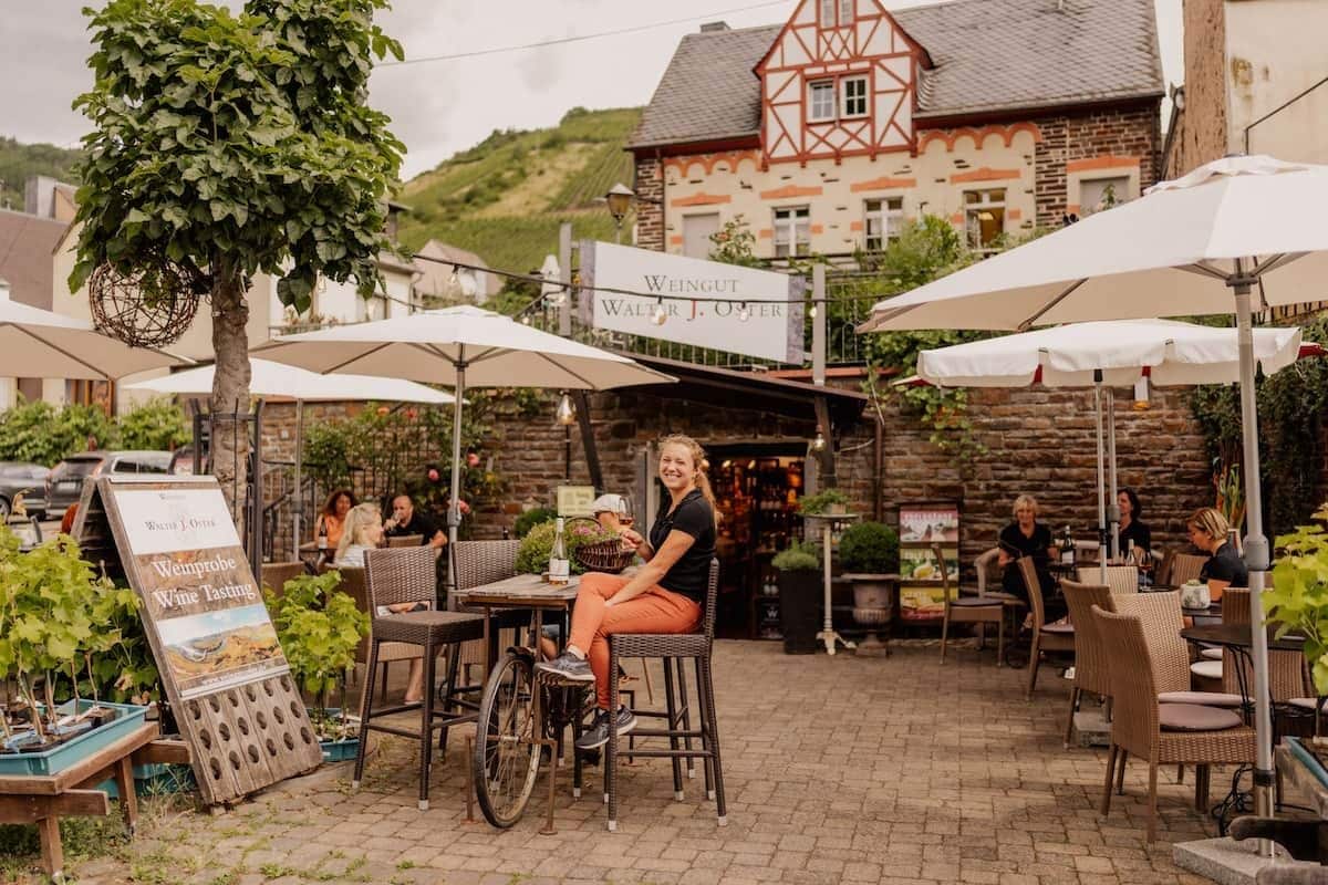 Un espace extérieur aménagé avec des chaises en osier et des parasols, une pancarte indiquant qu’il est possible de déguster du vin, et un bâtiment à l’arrière-plan dont la façade est de style traditionnel.