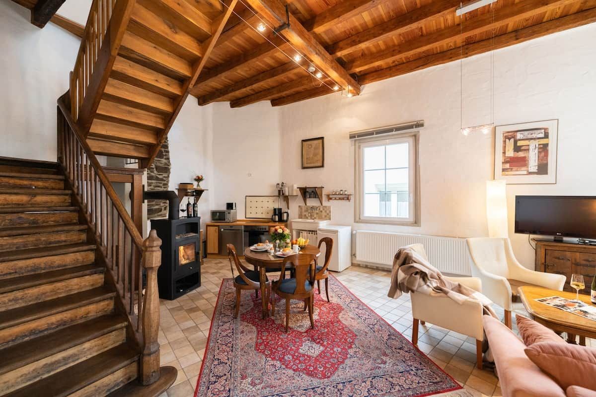 A cozy living room with a wooden staircase, a dining area with a red patterned rug, and a television.