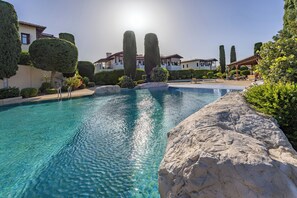 A clear swimming pool with a rock edge, surrounded by well-maintained landscaping and a sunny sky. - Zc02 Adjacent to Aphrodite Hills- Communal Pool, Garden (Kouklia)