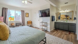 Quarto com cama, ventoinha no teto, televisão, penteadeira e janela com cortinas.