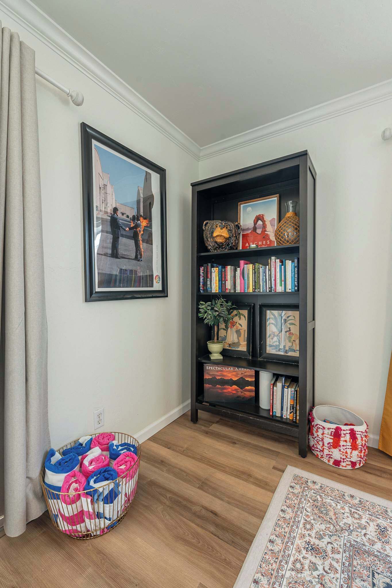 Ein Zimmer mit einem schwarzen Bücherregal, einem gerahmten Bild, einem farbenfrohen Ottomanen und einem gemusterten Teppich.