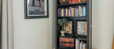 Ein Zimmer mit einem schwarzen Bücherregal, einem gerahmten Bild, einem farbenfrohen Ottomanen und einem gemusterten Teppich.