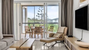A modern living room with a sofa, a lounge chair, a coffee table, and a TV. There is a balcony with a table and chairs, and a view of greenery and buildings. - Tropical Park View Retreat – 3BR Bali Resort in Kingscliff (1210) (Tweed)